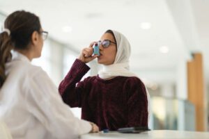 A Muslim woman wearing a hijab sits at a table while at a medical consultation. She has asthma and is learning to use an inhaler. A female doctor is watching and instructing the patient on how to use a puffer. The doctor's back is to the camera.